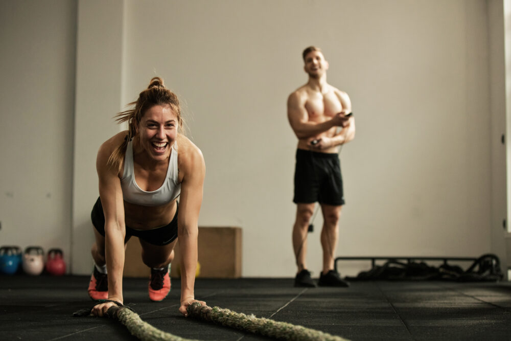 cheerful female athlete doing pushups while exercising with battle rope having fun gym there is man background